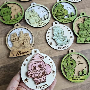 Può includere: Una collezione di decorazioni natalizie in legno con vari personaggi dei cartoni animati e nomi. Gli ornamenti presentano disegni tagliati al laser nei toni del verde, marrone e bianco. Ogni ornamento ha un anello per appenderlo e include il testo "Merry Christmas!"