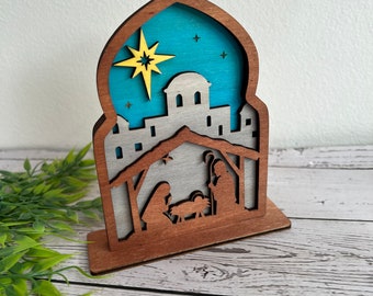 Pode incluir: Um presépio de madeira com um design em camadas. A cena apresenta uma estrela, uma cidade e silhuetas de Maria, José e o menino Jesus. O fundo é azul com detalhes de estrelas e cruzes. A base e a moldura são castanhas.
