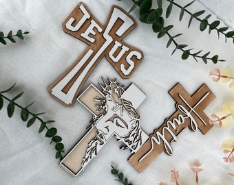 May include: Three wooden crosses with white painted designs. One cross features the word "Jesus" in white letters, another cross has a white silhouette of Jesus's face, and the third cross has the word "Joy" in white cursive writing.