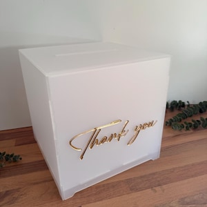 Puede incluir: Caja blanca de acrílico con ranura superior. La parte frontal de la caja tiene la inscripción dorada "Thank you". Diseñada para bodas, fiestas u otros eventos, para recoger tarjetas o notas.