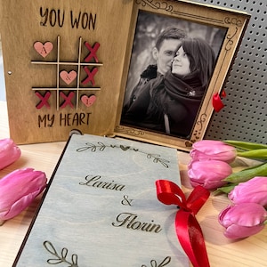 May include: A wooden box with a photo frame and a tic-tac-toe game on the front. The game says "You Won My Heart" and the photo frame has a black and white photo of a couple. The box is closed and has a red ribbon tied around it. The box is on a wooden surface with pink tulips.