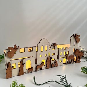 Puede incluir: Una escena de pueblo navideño de madera con una escena de la natividad, ángeles y palmeras. El pueblo está iluminado con luces blancas cálidas.