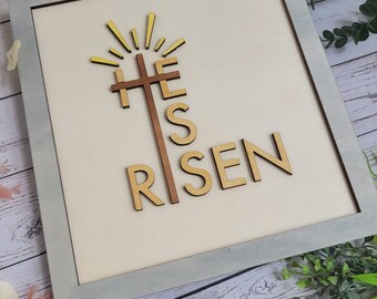 Pode incluir: Uma placa de madeira com uma moldura cinza clara e um fundo bege. A placa apresenta uma cruz de madeira com um sol radiante amarelo acima. As palavras "He is Risen" estão escritas em letras de madeira abaixo da cruz.
