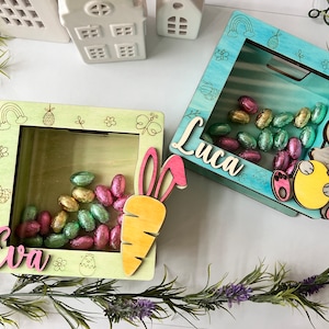 Op de afbeelding: Twee houten paasdozen met transparante kunststof ramen. De dozen zijn versierd met pastelkleuren en lentethema-ontwerpen. Eén doos heeft een wortel en de tekst "Eva" erop, de andere heeft een konijn en de tekst "Luca". Beide dozen zijn gevuld met chocolade-eieren.
