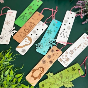 Può includere: Una collezione di segnalibri in legno con vari disegni a tema invernale. I segnalibri presentano fiocchi di neve, pinguini, alberi di Natale e agrifoglio. Ogni segnalibro ha un cordino rosso e bianco. La palette di colori include legno naturale, bianco, verde e blu.
