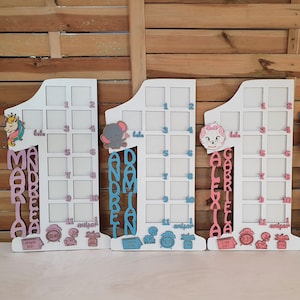 Può includere: Tre cornici portafoto in legno bianco a forma di numero uno. Ogni cornice ha ritagli per foto, un nome, elementi decorativi e la parola "babe". Le cornici sono per il primo anno di un bambino, con i nomi Maria, Andrei e Gabriela.