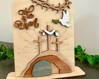 Pode incluir: Decoração de Páscoa em madeira com uma coroa de espinhos, uma pomba com um ramo de oliveira e três cruzes numa colina. A frase "He is Risen" está escrita. A decoração é feita de madeira em camadas com um acabamento natural.