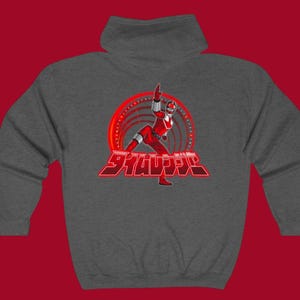 May include: A charcoal gray hooded sweatshirt featuring a red graphic of a costumed figure in a dynamic pose, set against a red circular design. Japanese text is below the figure. The sweatshirt has a high collar.