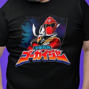 Peut inclure: T-shirt noir avec un graphisme coloré représentant un personnage rouge et or avec un casque, sur fond de drapeau déchiré. Du texte japonais est affiché en dessous du personnage. Le design est de style bande dessinée.