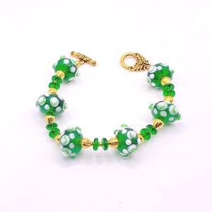 May include: A handmade bracelet featuring green glass beads with white polka dots, interspersed with gold-colored beads and smaller green beads. The bracelet has a gold-colored toggle clasp and a decorative hook clasp.