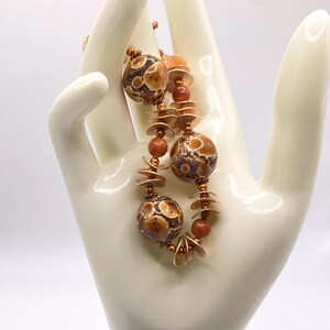 May include: A handcrafted necklace featuring large, speckled brown beads with gold accents, interspersed with smaller, round beads and flat, petal-shaped elements. The necklace has a warm, earthy color palette and a unique, artistic design.