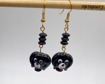 May include: A pair of gold-coloured dangle earrings. Each earring features a black dog-shaped charm with white and red accents. The earrings are suspended from gold-coloured fish hook ear wires and have a series of black beads.