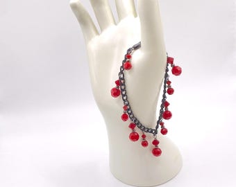 May include: A jewelry set featuring a dark gray chain bracelet and matching earrings, adorned with vibrant red, faceted beads. The bracelet is draped over a white hand-shaped display, with the earrings resting below.