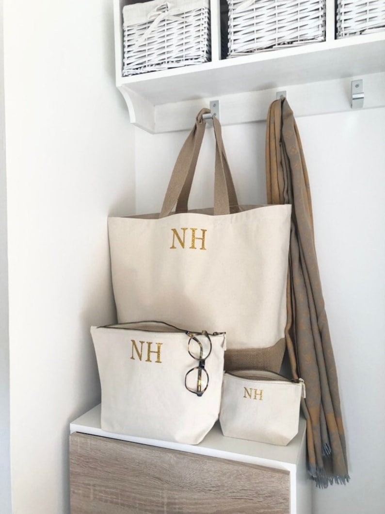 K&ouml;nnte beinhalten: Drei cremefarbene Canvas-Taschen mit goldenen Stickinitialen "NH" auf einem wei&szlig;en Regal. Die gr&ouml;&szlig;te Tasche ist eine Tragetasche mit braunen Jutegriffen. Die kleineren Taschen sind Rei&szlig;verschlusstaschen. Ein brauner Schal h&auml;ngt an einem Haken &uuml;ber den Taschen.