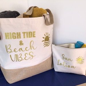 May include: A white canvas tote bag with a gold glitter design that says "High Tide & Beach Vibes". A smaller white canvas pouch with a gold glitter design that says "Sun Lotion" is in front of the tote bag.