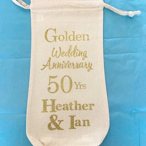 May include: A white cotton drawstring bag with gold glitter lettering that reads "Golden Wedding Anniversary 50 Yrs Heather & Ian"