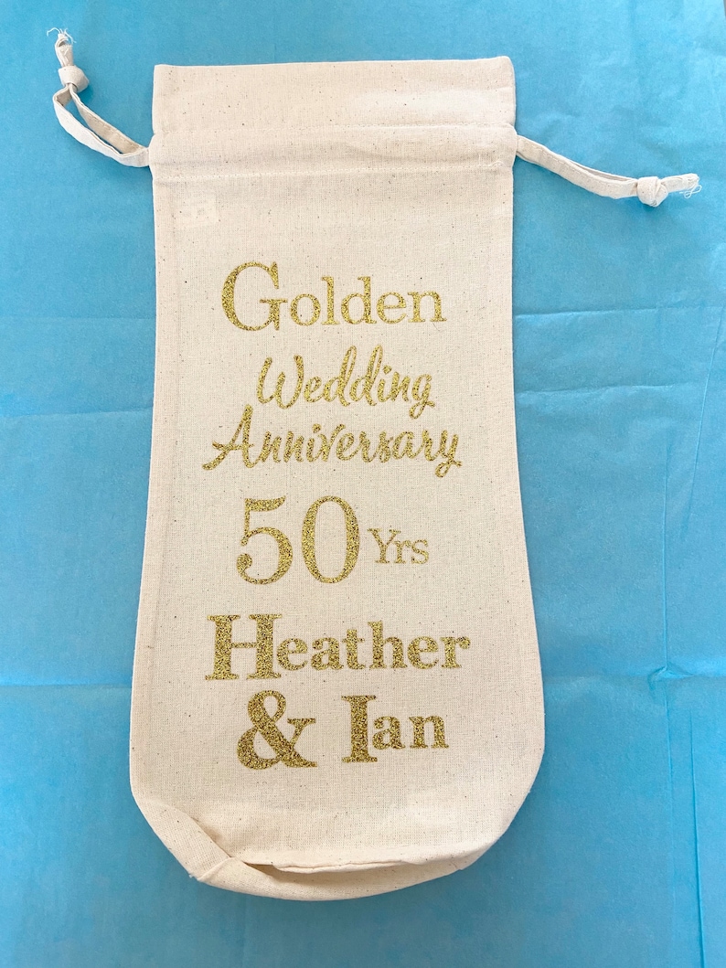 May include: A white cotton drawstring bag with gold glitter lettering that reads "Golden Wedding Anniversary 50 Yrs Heather & Ian"