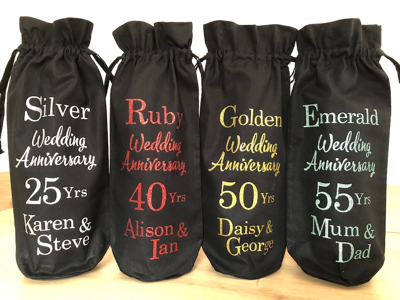 May include: Four black fabric wine bottle bags with different colored text. The bags are labeled with the text "Silver Wedding Anniversary 25 Yrs Karen & Steve", "Ruby Wedding Anniversary 40 Yrs Alison & Ian", "Golden Wedding Anniversary 50 Yrs Daisy & George", and "Emerald Wedding Anniversary 55 Yrs Mum & Dad".
