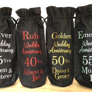 May include: Four black fabric wine bottle bags with different colored glitter text. The bags are labeled with the text "Silver Wedding Anniversary 25 Yrs Karen & Steve", "Ruby Wedding Anniversary 40 Yrs Alison & Ian", "Golden Wedding Anniversary 50 Yrs Daisy & George", and "Emerald Wedding Anniversary 55 Yrs Mum & Dad".
