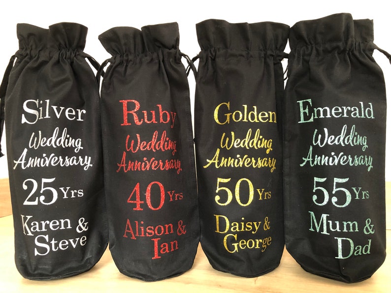 May include: Four black fabric wine bottle bags with different colored glitter text. The bags are labeled with the text "Silver Wedding Anniversary 25 Yrs Karen & Steve", "Ruby Wedding Anniversary 40 Yrs Alison & Ian", "Golden Wedding Anniversary 50 Yrs Daisy & George", and "Emerald Wedding Anniversary 55 Yrs Mum & Dad".