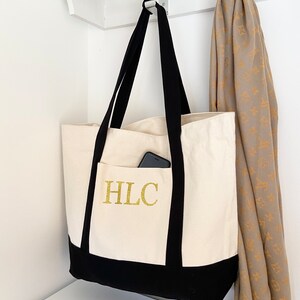 K&ouml;nnte beinhalten: Eine wei&szlig;e Canvas-Tragetasche mit schwarzen Henkeln und einem schwarzen Boden. Die Tasche hat ein golden gesticktes Monogramm "HLC" auf der Vorderseite. Ein Handy ist in der vorderen Tasche sichtbar.