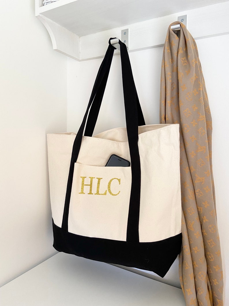 K&ouml;nnte beinhalten: Eine wei&szlig;e Canvas-Tragetasche mit schwarzen Henkeln und einem schwarzen Boden. Die Tasche hat ein golden gesticktes Monogramm "HLC" auf der Vorderseite. Ein Handy ist in der vorderen Tasche sichtbar.