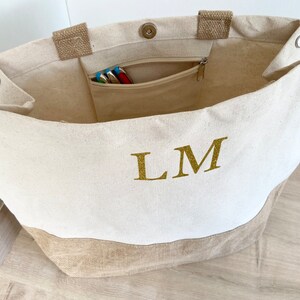 K&ouml;nnte beinhalten: Eine wei&szlig;e Canvas-Tragetasche mit braunem Jute-Boden. Die Tasche hat eine Rei&szlig;verschlusstasche innen und die Initialen "LM" sind in goldenen Glitzer auf der Vorderseite gestickt.