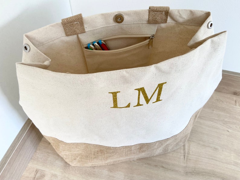 K&ouml;nnte beinhalten: Eine wei&szlig;e Canvas-Tragetasche mit braunem Jute-Boden. Die Tasche hat eine Rei&szlig;verschlusstasche innen und die Initialen "LM" sind in goldenen Glitzer auf der Vorderseite gestickt.