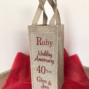 May include: A beige linen wine bottle gift bag with red embroidered text that reads "Ruby Wedding Anniversary 40 Yrs Glen & Jan".