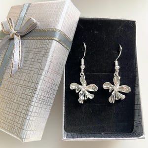 May include: A pair of silver-toned drop earrings featuring a stylised, multi-petaled flower design with a small clear stone at the centre of each. The earrings hang from silver-toned hooks and are presented inside a rectangular gift box.