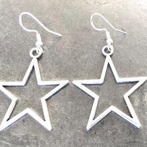 May include: A pair of silver-toned earrings featuring open-frame star pendants. Each star is suspended from a simple silver-toned hook wire, creating a dangling design. The metal has a smooth, reflective finish, showcasing a clean, geometric star shape.