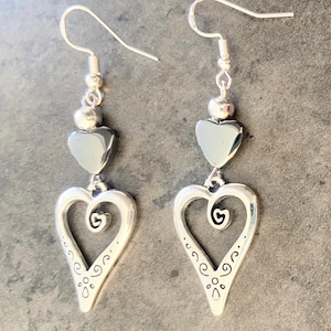 May include: A pair of silver-toned drop earrings featuring a polished dark grey heart bead suspended above a larger openwork heart charm. The lower heart charm displays a central spiral detail and small etched dots along the bottom edges.
