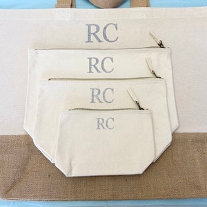 K&ouml;nnte beinhalten: Ein Set aus vier Canvas-Taschen mit Rei&szlig;verschl&uuml;ssen. Die Taschen sind ineinander verschachtelt, wobei die gr&ouml;&szlig;te Tasche unten und die kleinste Tasche oben liegt. Jede Tasche hat die Initialen "RC" in silberner Glitzer-Schrift auf der Vorderseite gestickt.