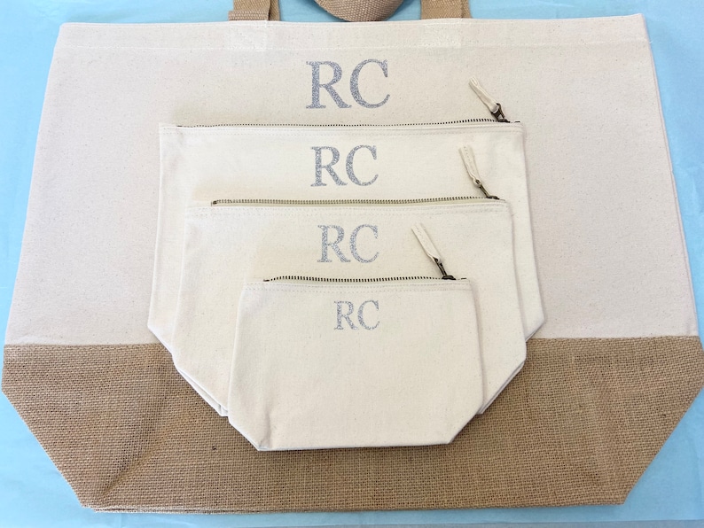 K&ouml;nnte beinhalten: Ein Set aus vier Canvas-Taschen mit Rei&szlig;verschl&uuml;ssen. Die Taschen sind ineinander verschachtelt, wobei die gr&ouml;&szlig;te Tasche unten und die kleinste Tasche oben liegt. Jede Tasche hat die Initialen "RC" in silberner Glitzer-Schrift auf der Vorderseite gestickt.