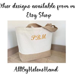 K&ouml;nnte beinhalten: Eine wei&szlig;e Canvas-Tragetasche mit braunem Jutebesatz und Griffen. Die Tasche hat die Initialen "P.B.M." in Orange auf der Vorderseite gestickt. Die Tasche steht auf einer braunen Oberfl&auml;che.