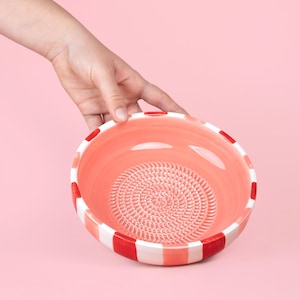 Könnte beinhalten: Eine runde Reibeschale aus Keramik mit korallenrosa Innenseite und einem strukturierten Spiralmuster am Boden zum Reiben. Die Außenseite der Schale ist mit abwechselnden vertikalen Streifen in Weiß und leuchtendem Rot verziert, was ein auffälliges Muster ergibt.