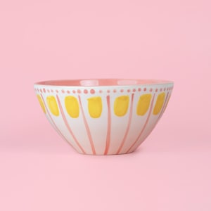 May include: A white ceramic bowl with a pink interior and a decorative exterior. The exterior features vertical pink lines with yellow oblong shapes and pink dots. The bowl is set against a pink background.