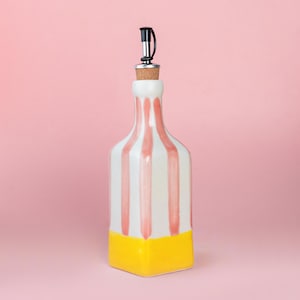 Könnte beinhalten: Eine dekorative Keramikflasche mit schwarzem und silbernem Ausgießer. Die Flasche ist weiß mit vertikalen rosa Streifen und einem gelben Boden. Die Flasche ist für Flüssigkeiten konzipiert.