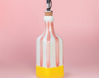 May include: A decorative ceramic bottle with a black and silver spout. The bottle is white with vertical pink stripes and a yellow base. The bottle is designed for holding liquids.