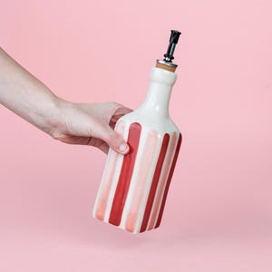 May include: A white ceramic bottle with vertical red and pink stripes, held by a hand. The bottle has a black spout and a cork stopper. The background is a solid pink.