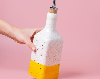 May include: A white ceramic bottle with a speckled design and a bright yellow base. It has a metal spout and a cork stopper. The bottle is held by a hand against a pink background. This decorative bottle is perfect for oils and vinegars.