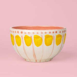Może przedstawiać: Ceramiczna miska z białym tłem, ozdobiona ręcznie malowanym wzorem w pionowe różowe paski oraz jasne, żółte owalne pociągnięcia pędzla. Krawędź jest udekorowana rzędem różowych kropek, a wnętrze miski ma jednolity jasnoróżowy kolor.