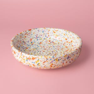 Puede incluir: Un cuenco rallador de cerámica redondo con base blanca y un patrón de motas coloridas en tonos naranja, azul, amarillo y morado. El fondo interior del cuenco presenta un patrón circular texturizado en relieve diseñado para rallar alimentos.