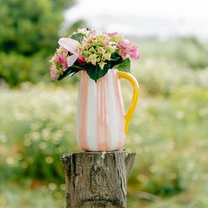 Puede incluir: Una jarra de cerámica con rayas verticales rosas y blancas y un asa amarilla, llena de flores rosas y verdes. La jarra está sobre un poste de madera desgastada, con un fondo verde borroso.