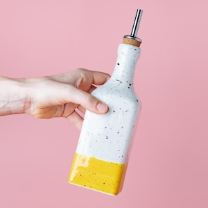 Puede incluir: Una botella de cerámica blanca con manchas amarillas y una mitad inferior amarilla. La botella tiene un pico de vertido plateado y un tapón de corcho. La botella está siendo sostenida por una mano.