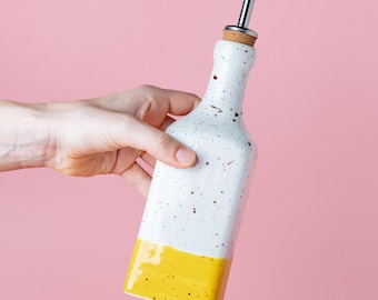 May include: A white ceramic bottle with a speckled finish and a bright yellow base. The bottle features a cork stopper topped with a silver-coloured metal pour spout. The design is rectangular with rounded edges, providing a functional container.