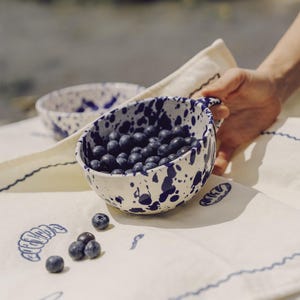 May include: A white ceramic bowl with a blue splatter design, filled with fresh blueberries. The bowl is held over a cream-colored linen cloth with blue embroidered food illustrations. Another bowl is in the background.