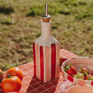 Può includere: Una bottiglia decorativa in ceramica con un design a righe rosse e bianche, sormontata da un tappo in sughero e un beccuccio in metallo. La bottiglia è su una tovaglia a scacchi rossi e bianchi, con pomodori e una ciotola di fragole.