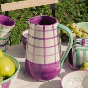 Peut inclure: Une carafe en céramique blanche avec un motif à carreaux violets et une poignée verte. La carafe fait partie d'un ensemble comprenant des bols et des tasses, tous avec des accents violets et verts assortis. L'ensemble est disposé sur une nappe blanche, avec des raisins et des pommes à proximité.