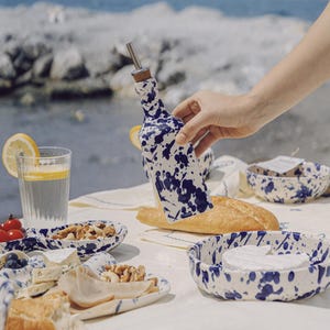 Puede incluir: Una botella de cerámica blanca con un diseño de salpicaduras azules, sostenida por una mano. La botella tiene un pico de metal y un tapón de corcho. La mesa incluye pan, queso, nueces y cuencos pequeños con el mismo patrón.